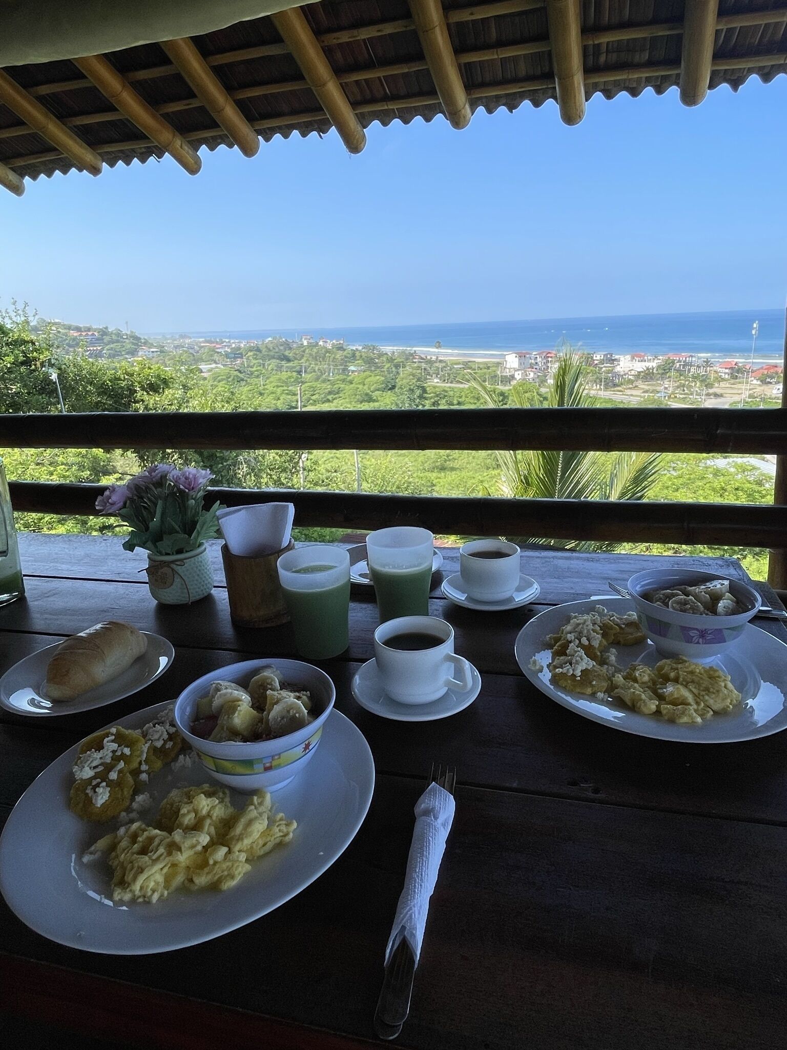 Una mesa dispuesta con alimentos para el desayuno, como huevos, pasteles y café, con vistas a un paisaje costero pintoresco.