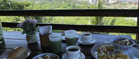 Una mesa dispuesta con alimentos para el desayuno, como huevos, pasteles y café, con vistas a un paisaje costero pintoresco.