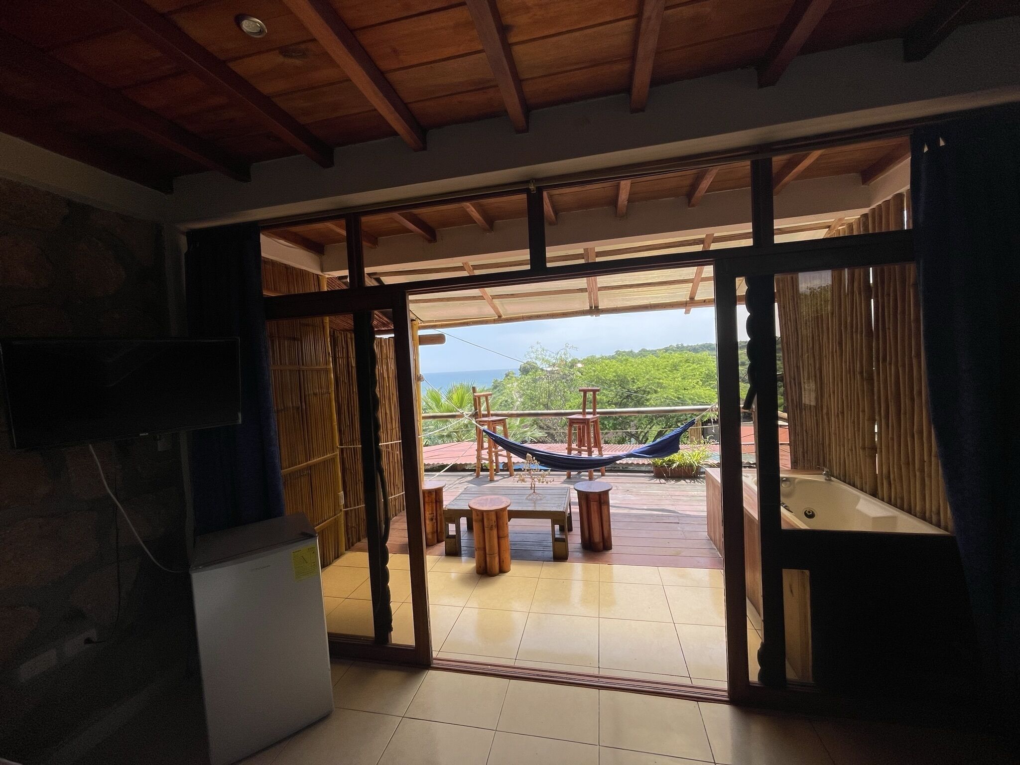 Un ventanal abierto que da a un balcón con hamaca, mobiliario de madera y vista al mar.
