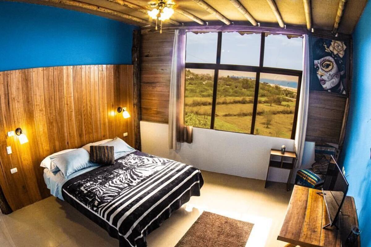 Une chambre à coucher avec une tête de lit en bois, un lit recouvert d’une couverture à motifs zébrés, un bureau avec une lampe et une grande fenêtre donnant sur un champ.