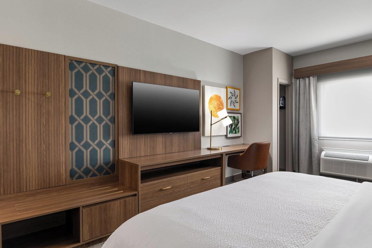 Une chambre d’hôtel avec un grand lit, un bureau en bois avec un téléviseur à écran plat, une chaise et une fenêtre avec des rideaux.
