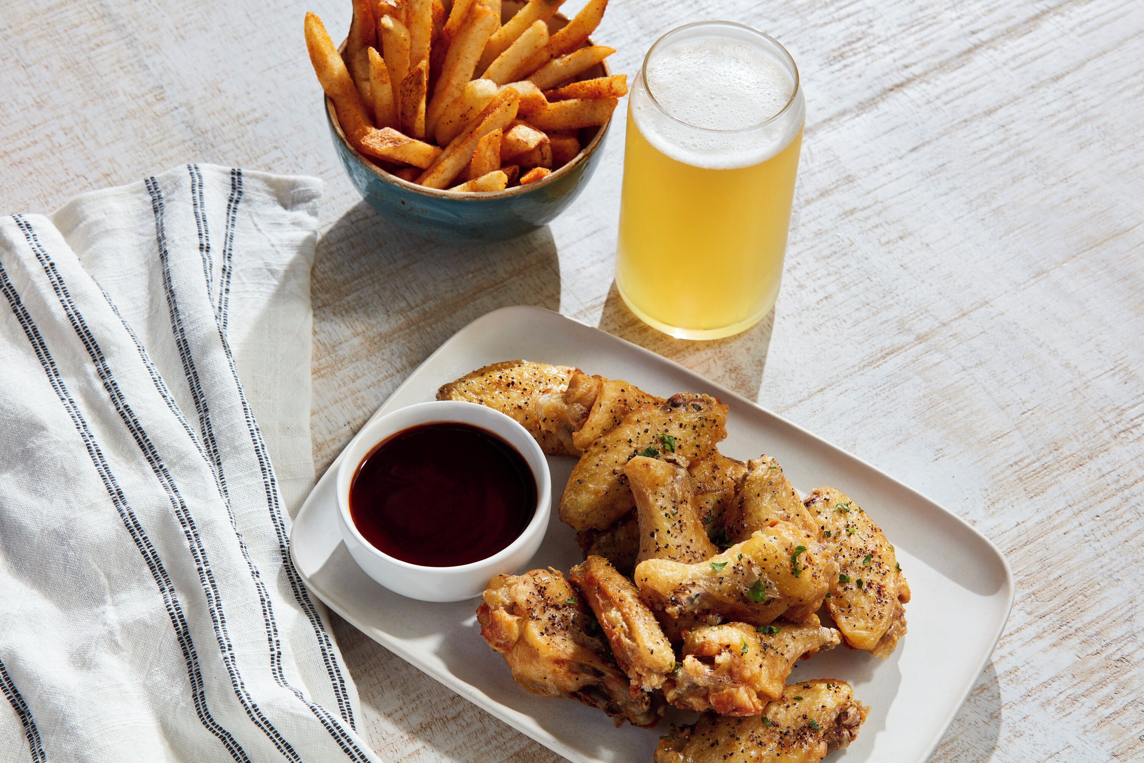A plate of fried chicken wings with dipping sauce, a bowl of fries, and a glass of beer on a wooden table.