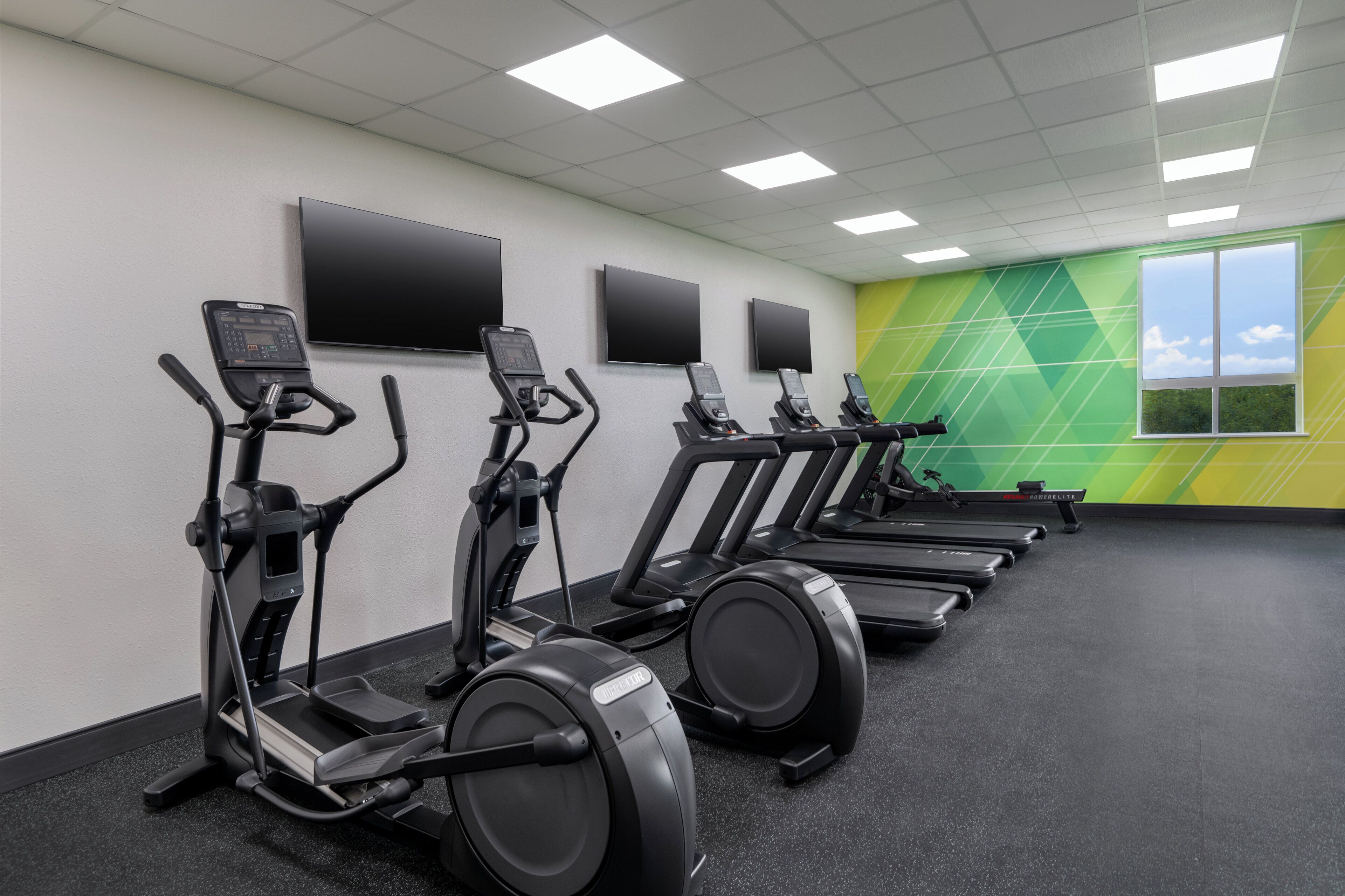 A gym with multiple treadmills and elliptical machines, a large TV screen, and a green wall with a window.