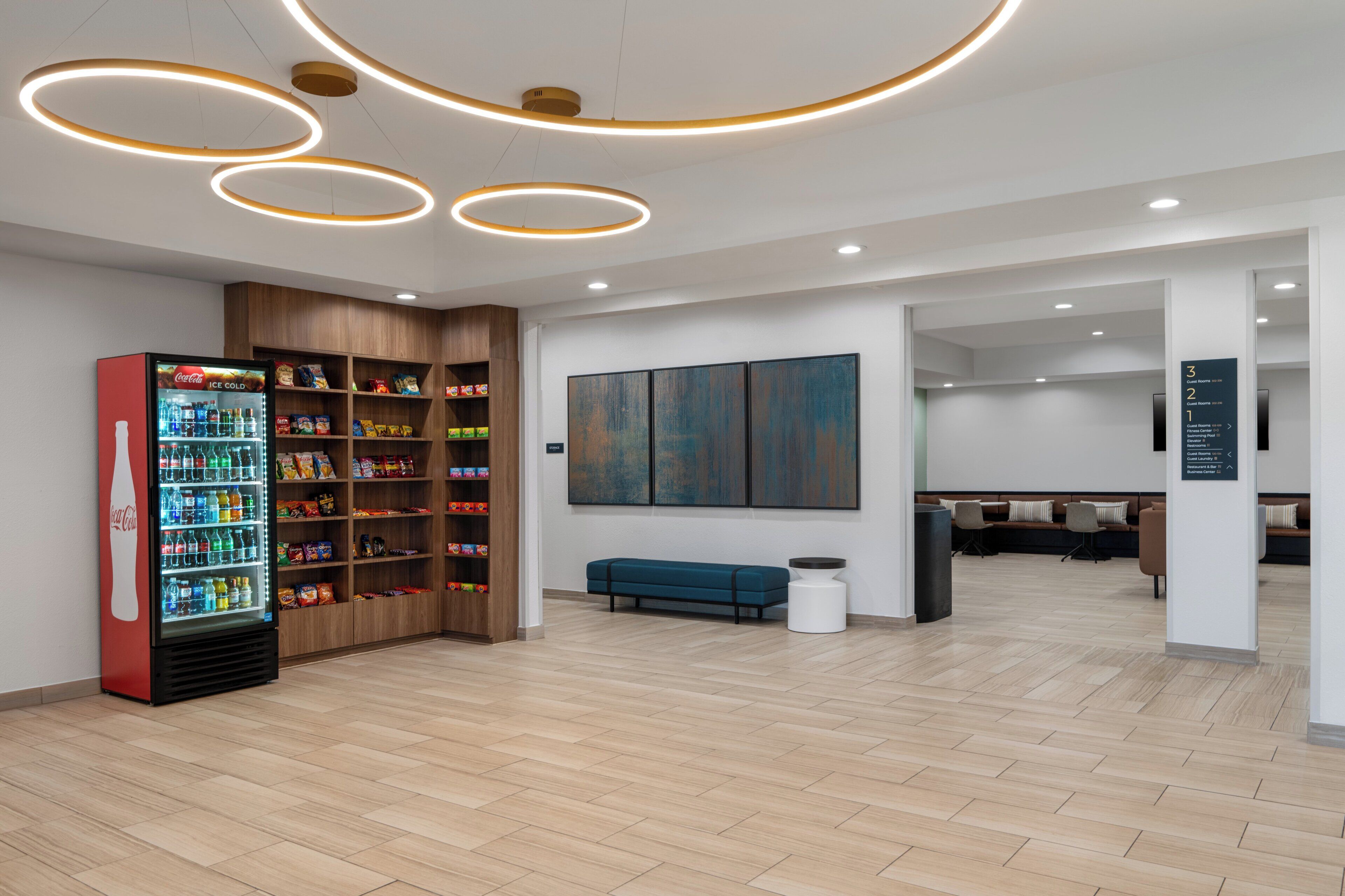 A modern lobby with a Coca-Cola refrigerator stocked with beverages, wooden shelving, a blue bench, and a large abstract painting on the wall.