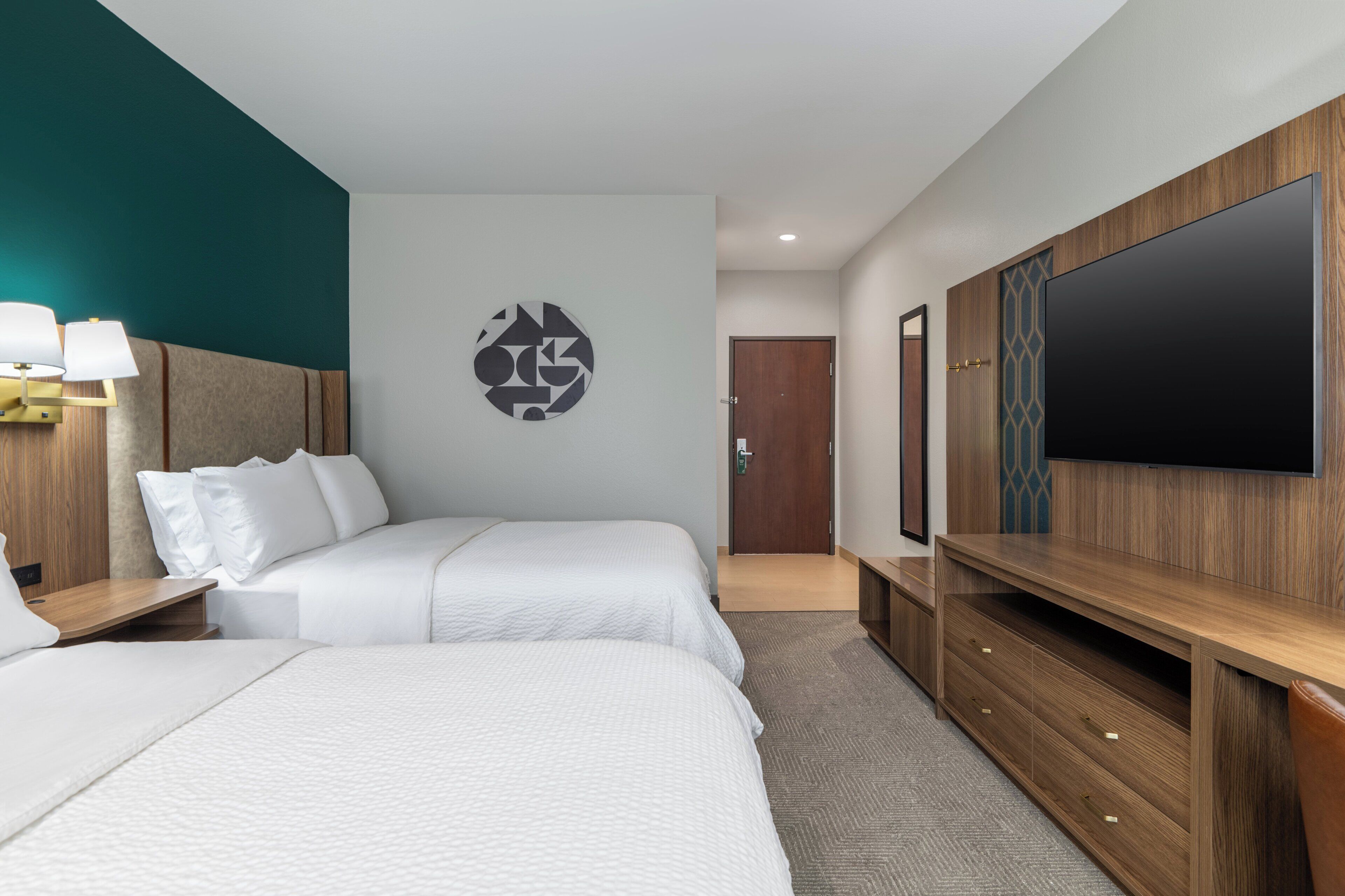 Ein Hotelzimmer mit zwei Betten, einem großen Flachbildfernseher, einer Holztruhe zur Unterhaltung und einem an der Wand befestigten Kunstwerk.
