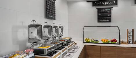 Zona de bufé con múltiples dispensadores de comida, una sección de frutas y yogur, y un cartel que dice 'conoce tu comida'.