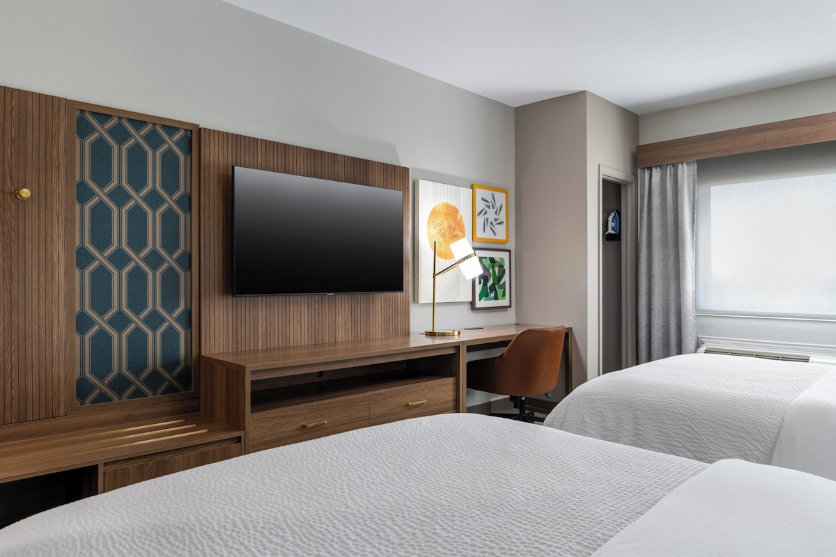 Une chambre d’hôtel avec deux lits, une télévision, un bureau avec une chaise et des œuvres d’art accrochées au mur.