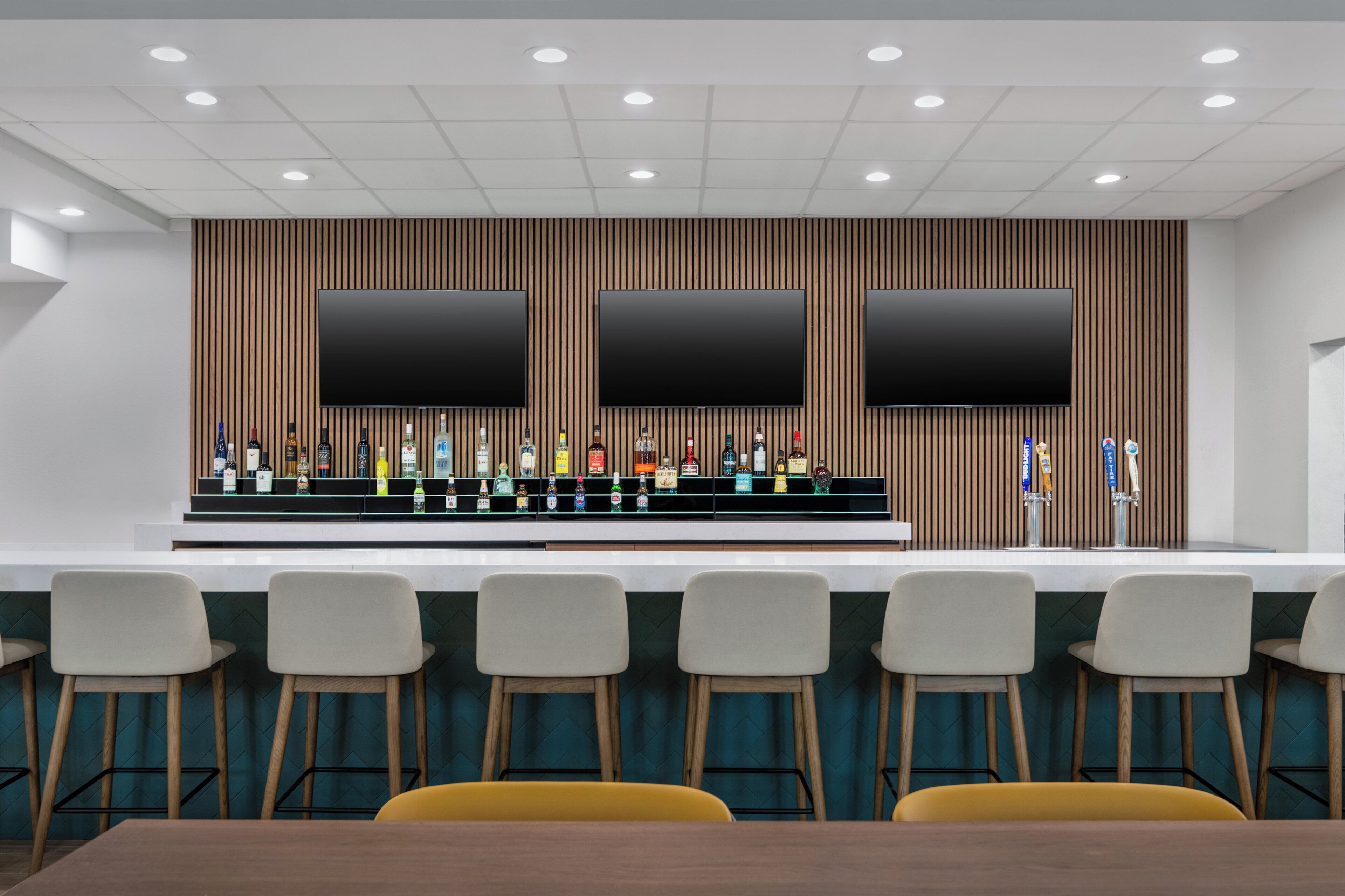 A bar area with three large screens, a variety of bottles, and bar stools.