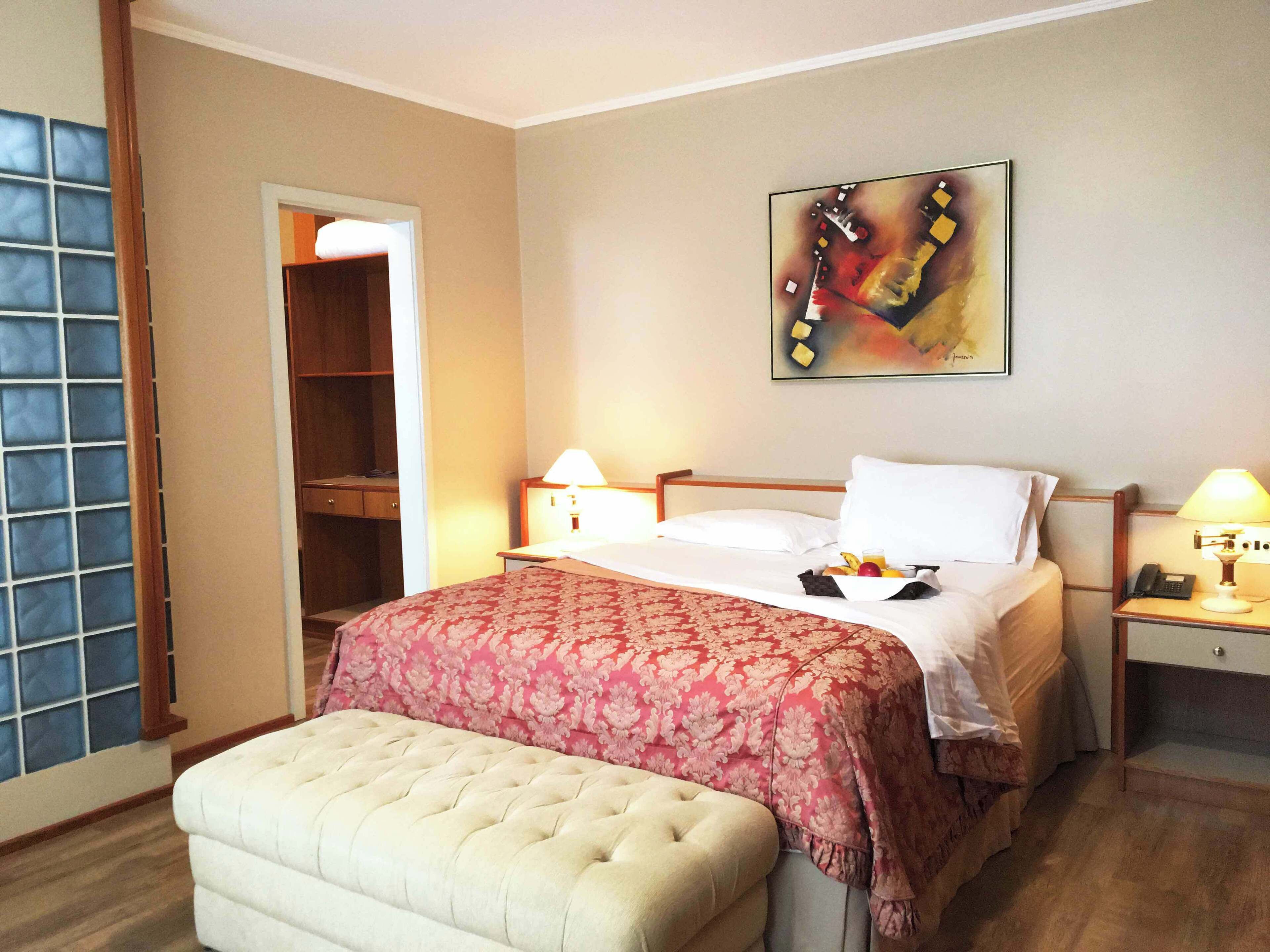 Habitación de hotel con cama, lámparas de noche, mesita de noche con teléfono, una pequeña mesa con fruta y una taza, un banco, un armario y un cuadro en la pared.
