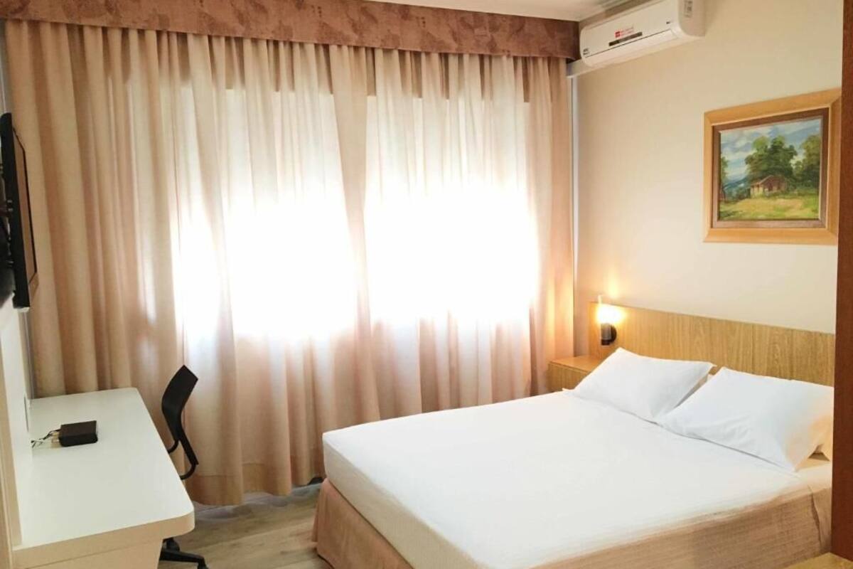 A hotel room with a bed, desk, chair, and a painting on the wall.