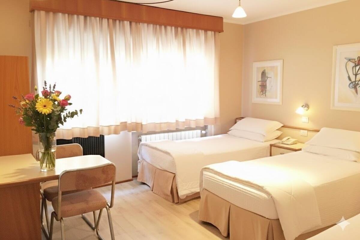 A hotel room with two beds, a dining table, chairs, a vase with flowers, and artwork on the walls.