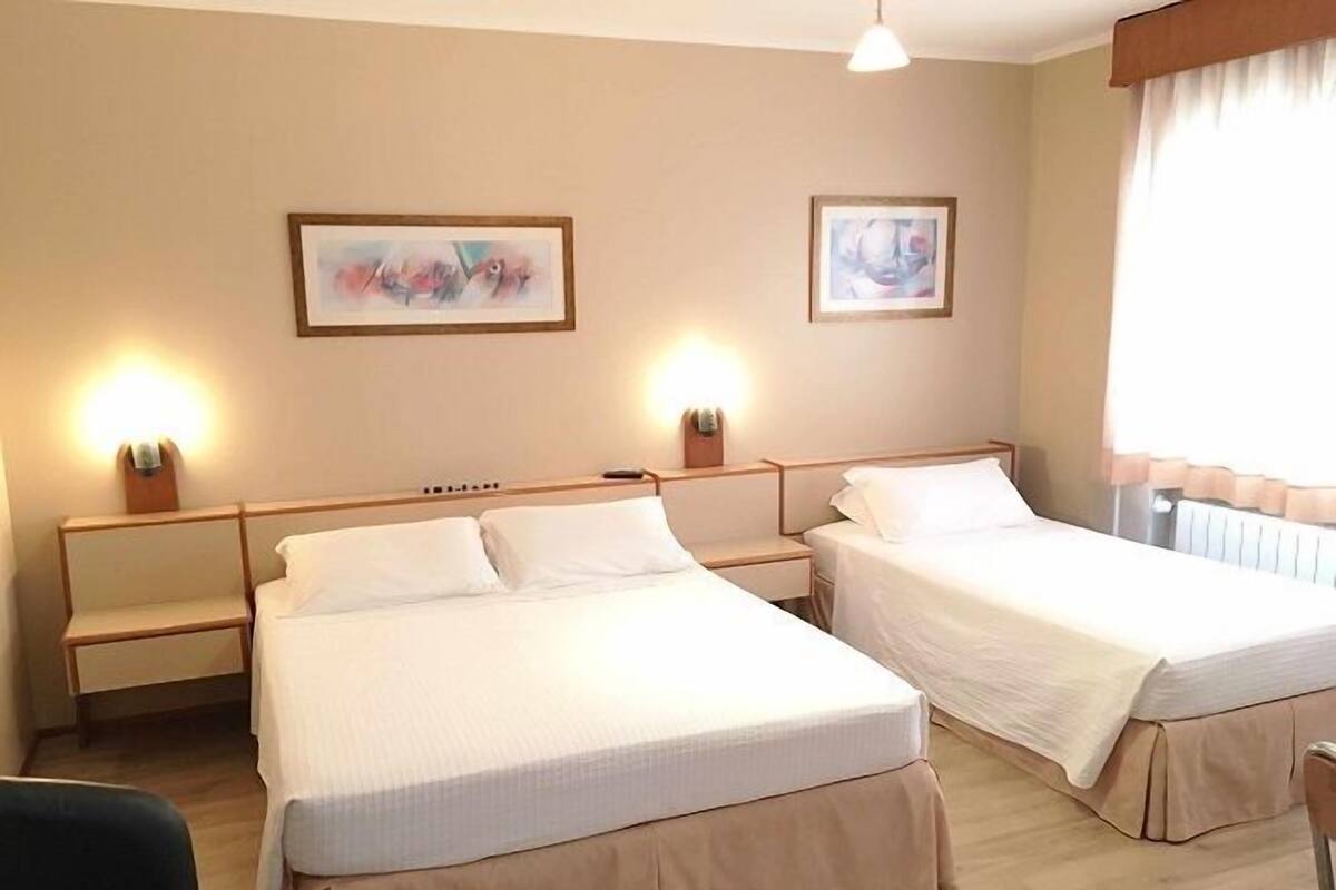 A hotel room with two single beds, a wooden headboard, a nightstand with a lamp, and two framed pictures on the wall.