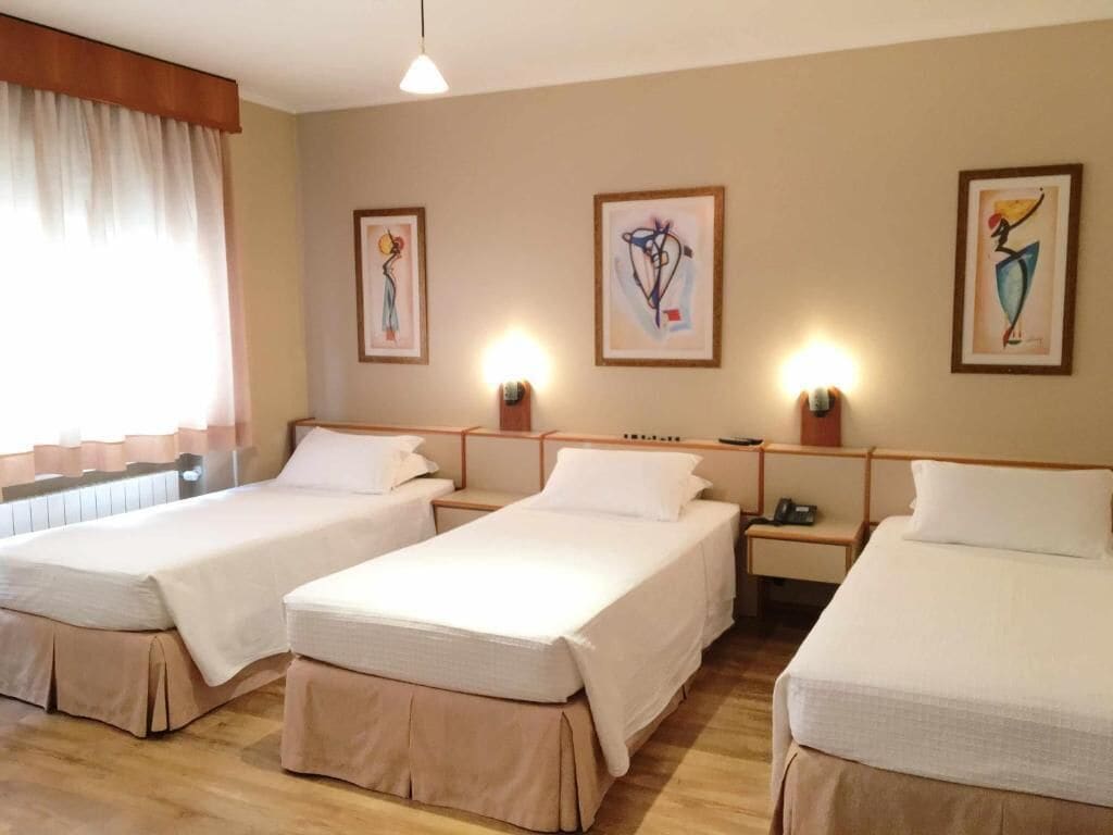 A hotel room with three single beds, wooden headboards, bedside tables, and artwork on the walls.