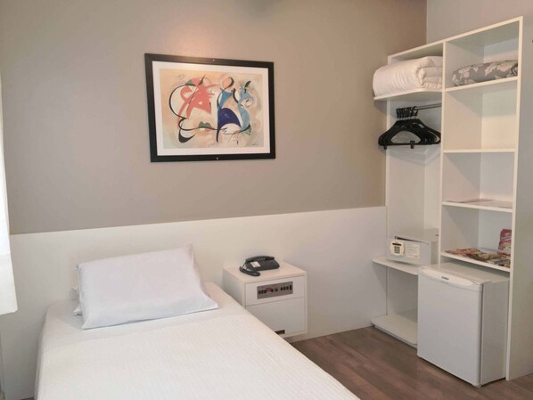 Une chambre à coucher comprenant un lit, une table de chevet avec un téléphone, un petit réfrigérateur et une armoire blanche avec des étagères.