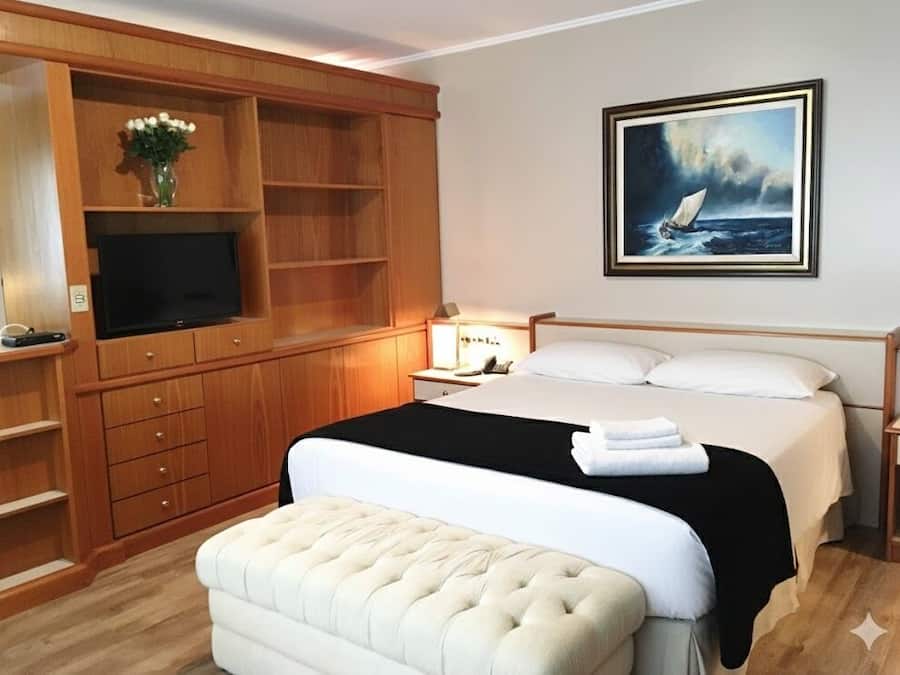 A hotel room with a large bed, a television, wooden built-in shelving, and a painting of a sailboat.