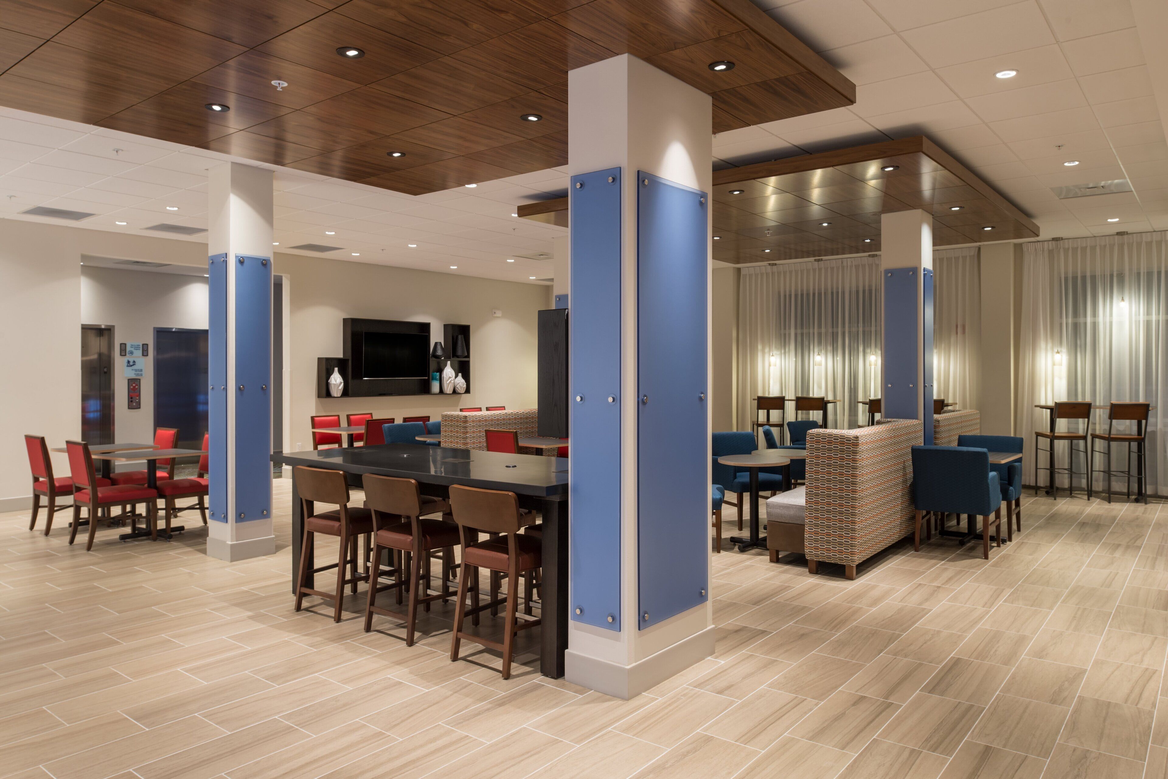 A modern hotel lobby with a central dining area, seating arrangements, and a television screen.