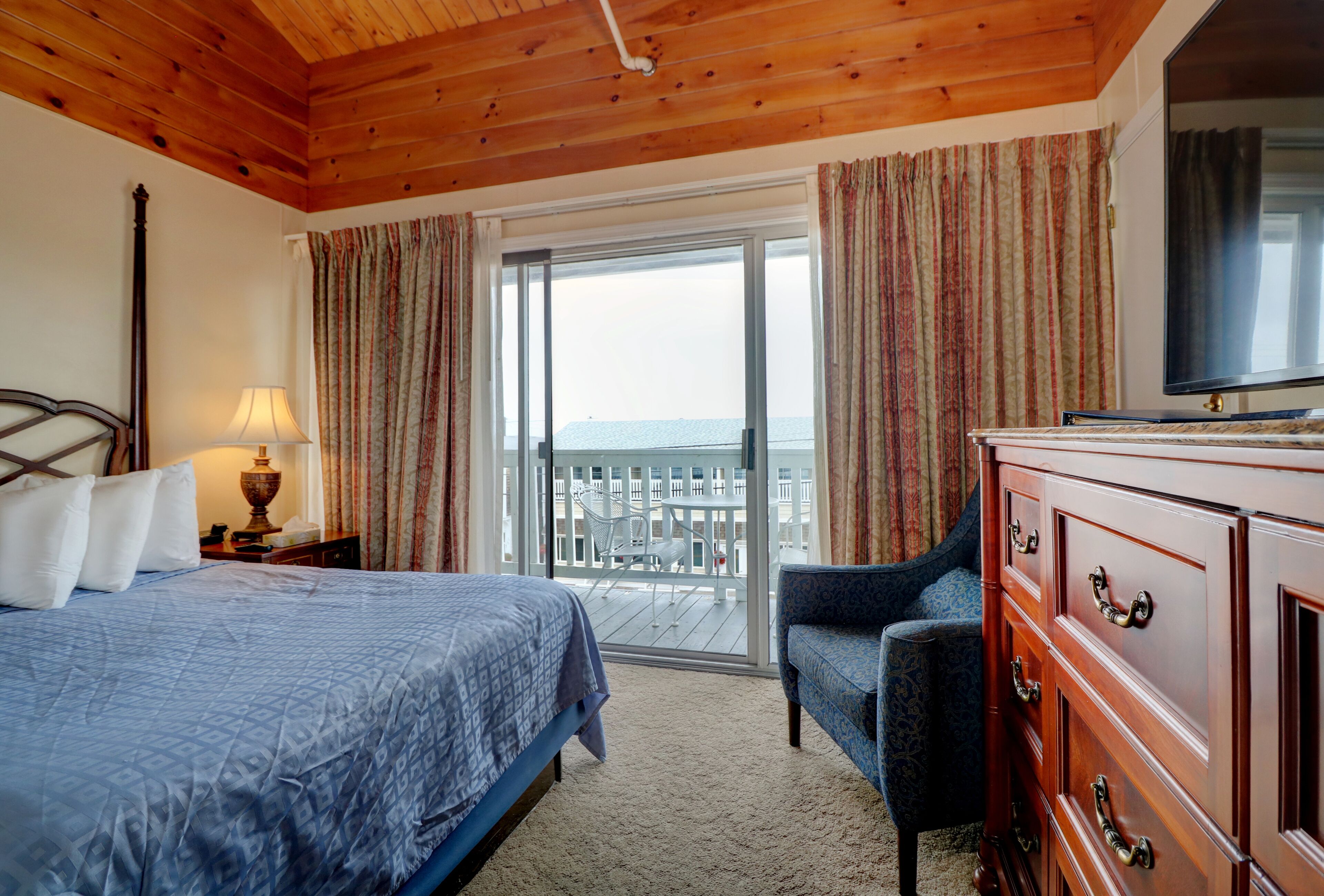 A hotel room with a bed, a dresser, a chair, a lamp, and a balcony with a view.