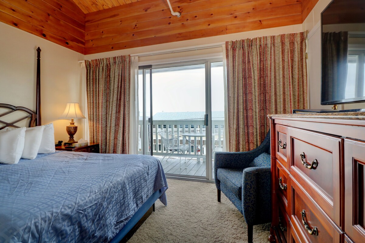 A hotel room with a bed, a dresser, a chair, a lamp, and a balcony with a view.