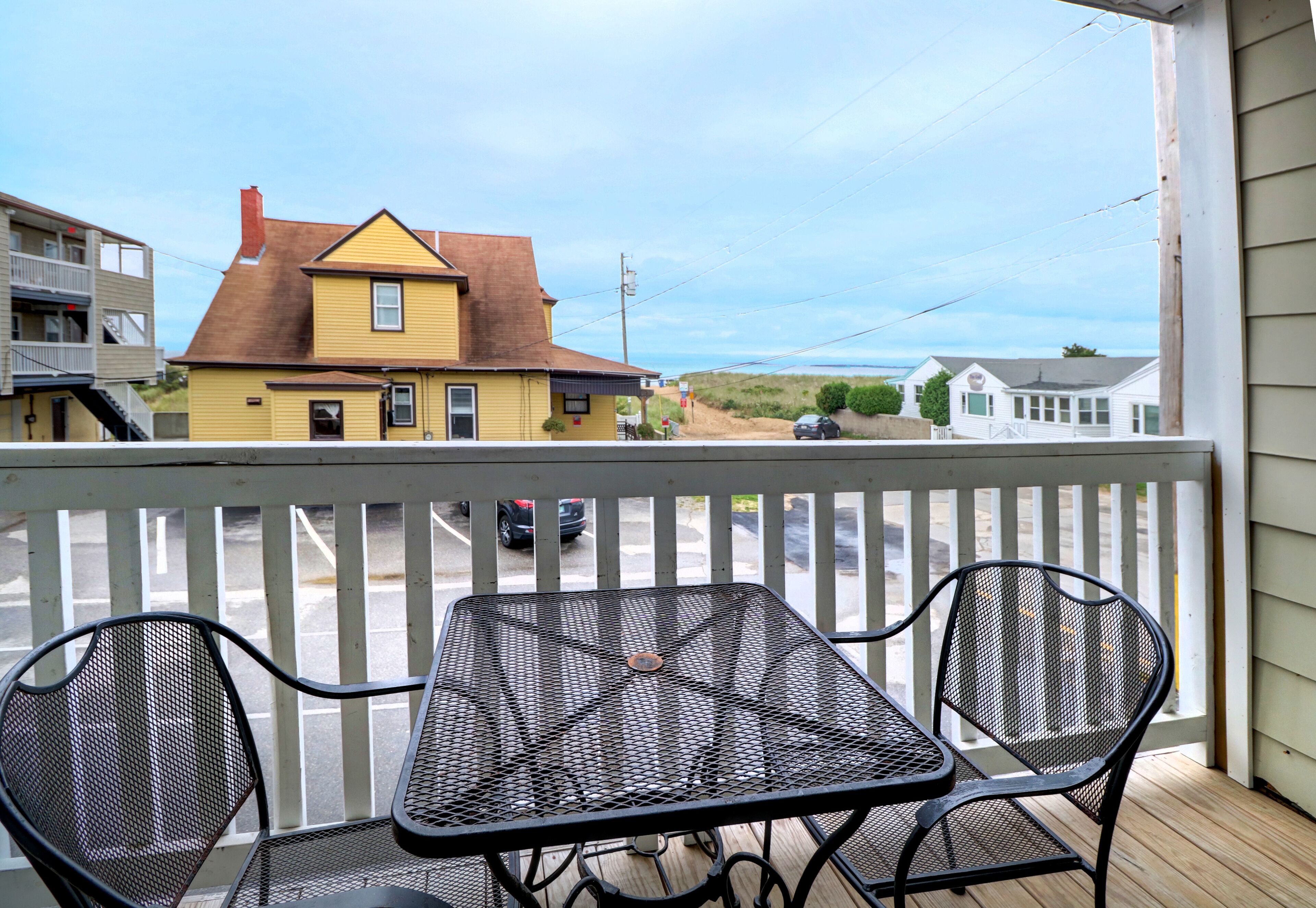 A balcony with a metal table and two chairs, overlooking a street with houses and a parked car.