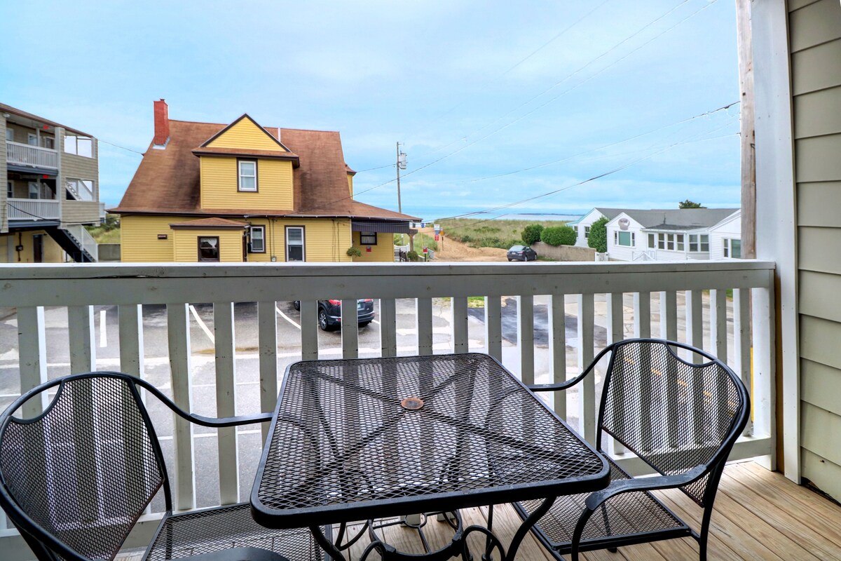 A balcony with a metal table and two chairs, overlooking a street with houses and a parked car.