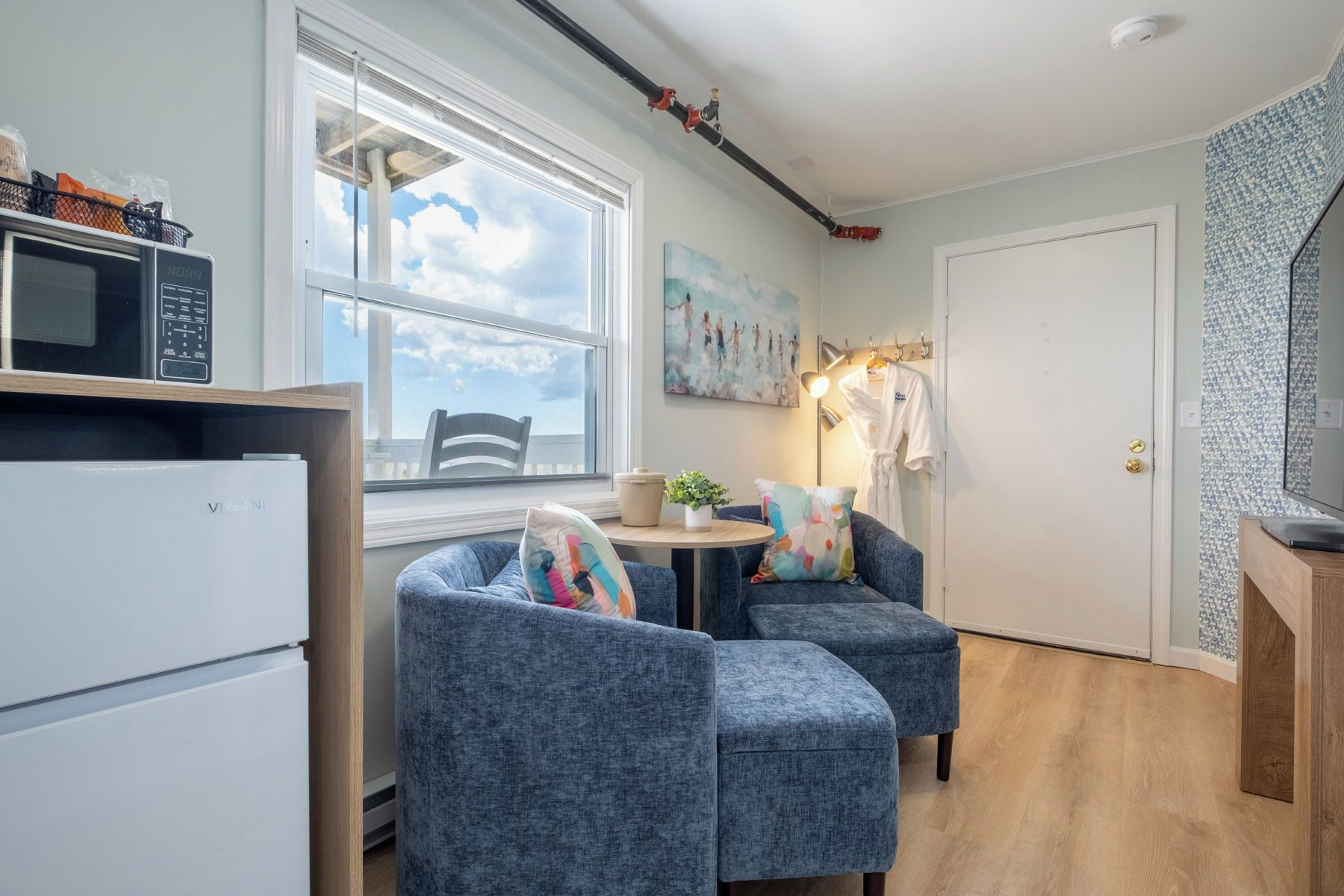 Ett kompakt bostadsutrymme med ett litet kök, en matplats med två blå stolar, ett litet runt bord och en tavla på väggen.