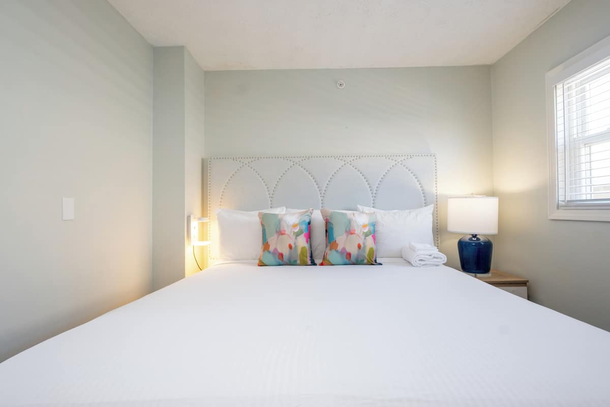 A hotel room with a large bed, a headboard with a decorative pattern, a bedside lamp, and a window with blinds.