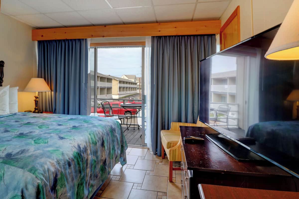 A hotel room with a bed, a desk, a television, and a view of a building and a parking area through a glass door.