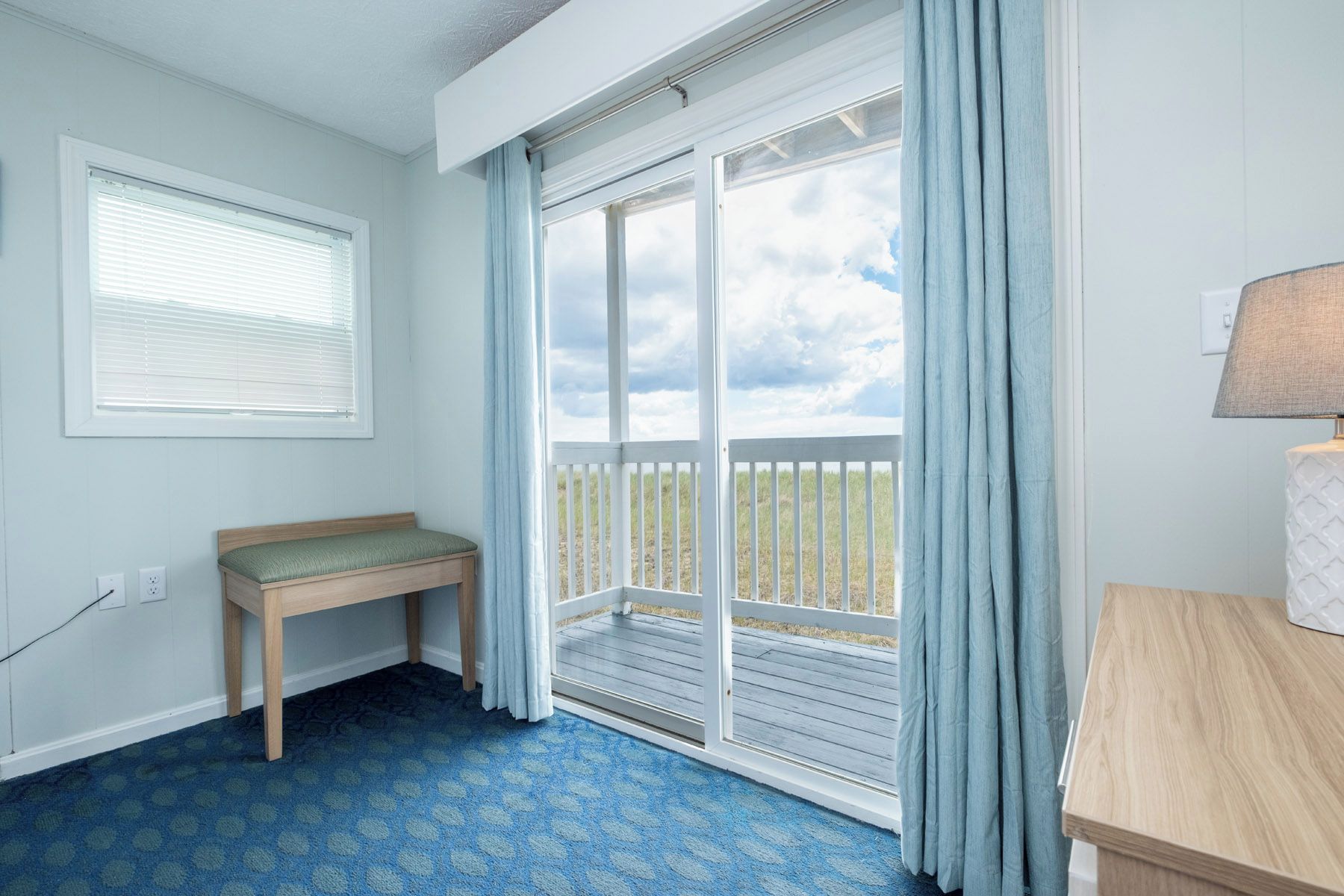 A room with a bench, a table, a window with blinds, and a view of the outdoors through a sliding door.