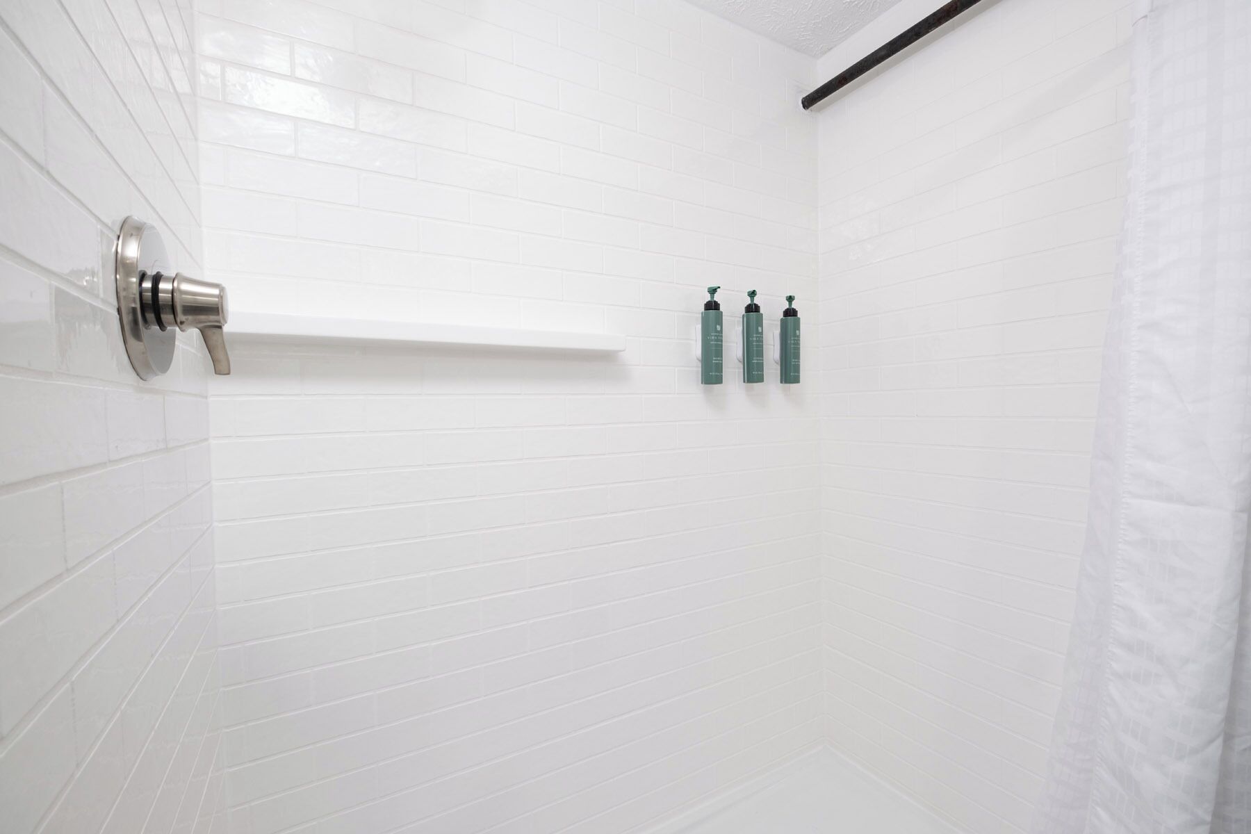 A white-tiled shower with a faucet and three green bottles on the wall.