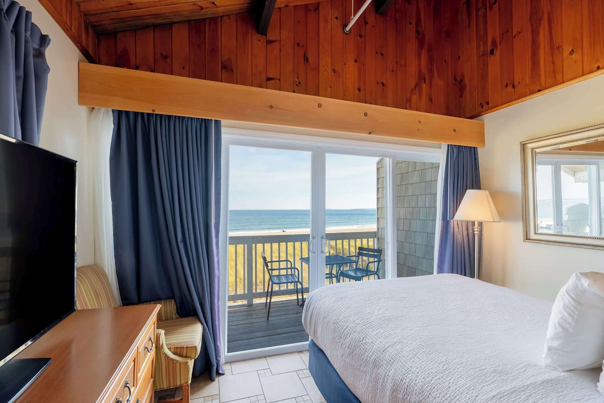 A hotel room with a wooden ceiling, a bed, a TV, a desk, and a balcony with a view of the sea.