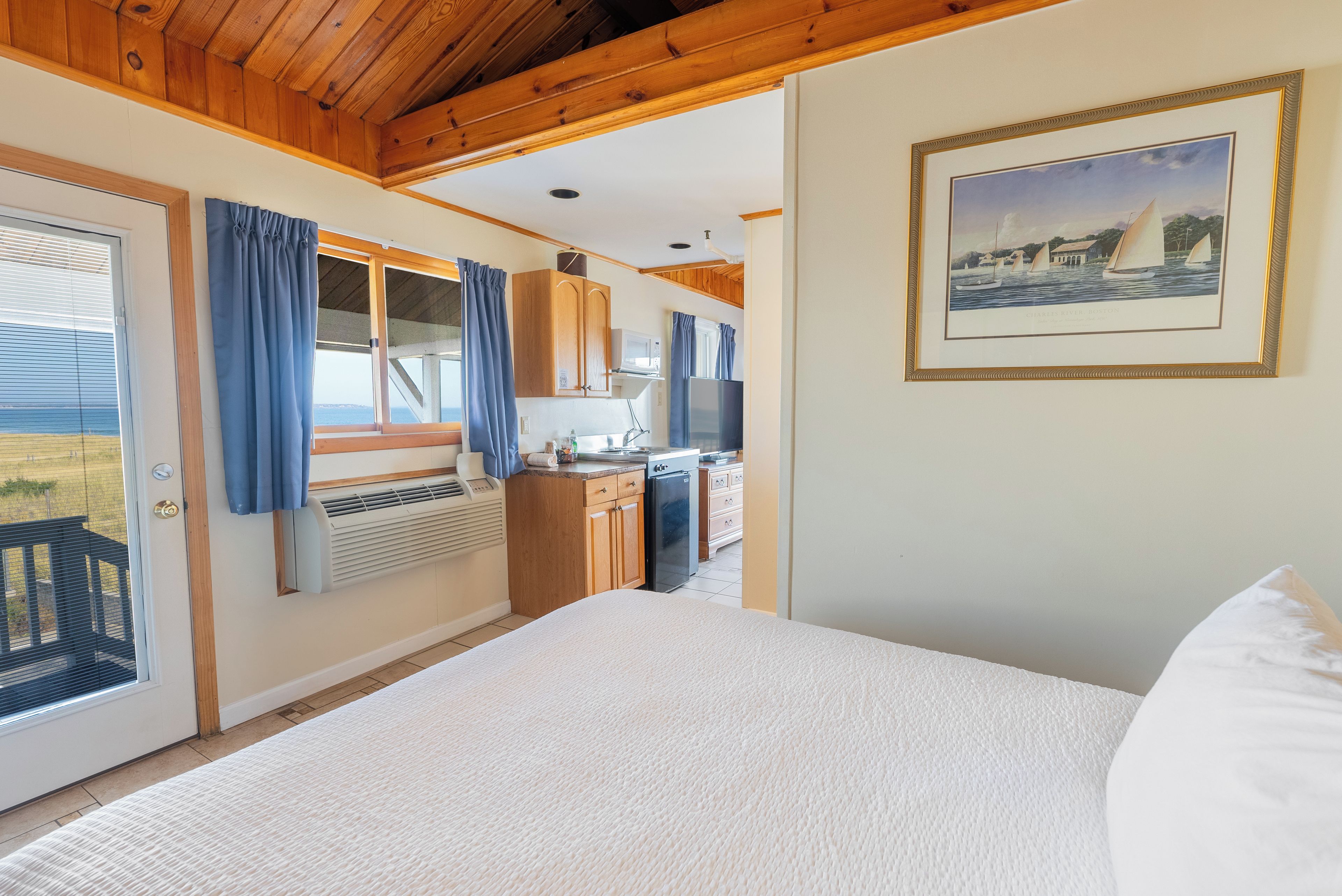 A bedroom with a large bed, a view of the kitchen, and a framed painting on the wall.