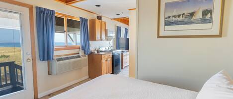 A bedroom with a large bed, a view of the kitchen, and a framed painting on the wall.