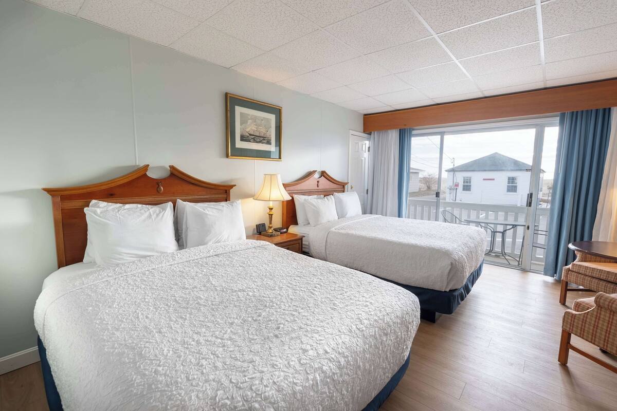 A hotel room with two beds, a nightstand with a lamp, a framed picture on the wall, and a view of the outside through a sliding door.