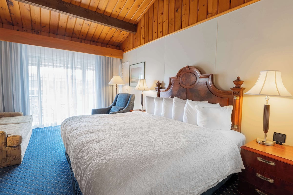 A hotel room with a large bed, a chair, a desk with a lamp, and a window with curtains.