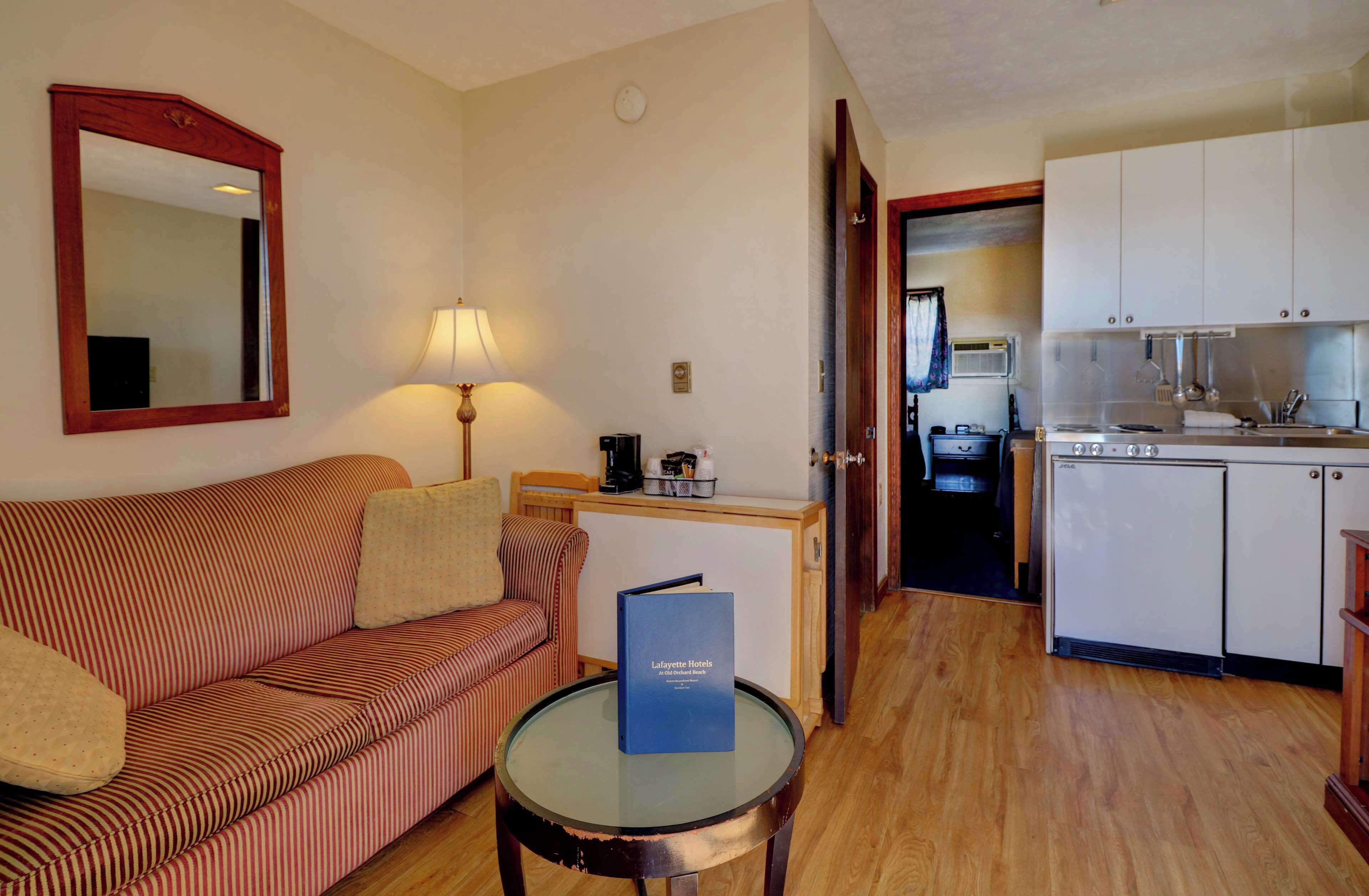 A compact hotel room with a striped sofa, a small table with a book, a lamp, and a kitchenette with a stove and sink.