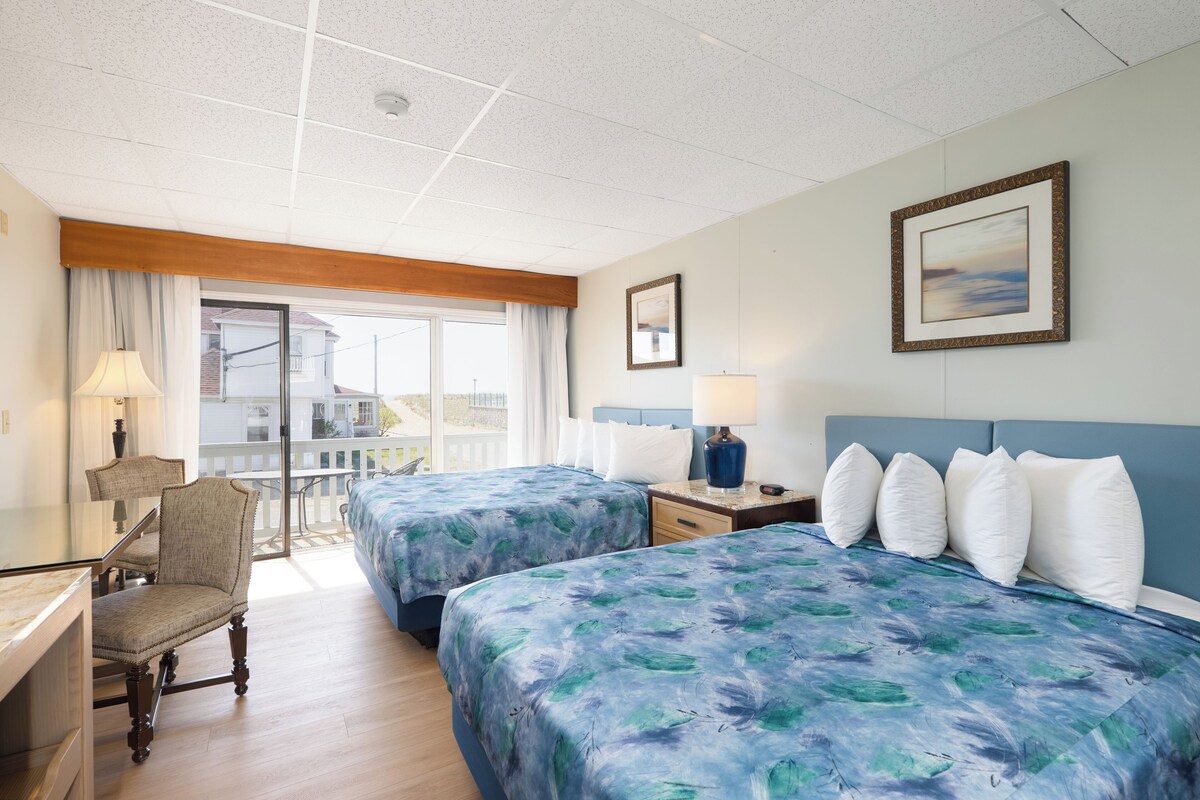 A hotel room with two beds, a desk, a chair, a lamp, and a window with a view.