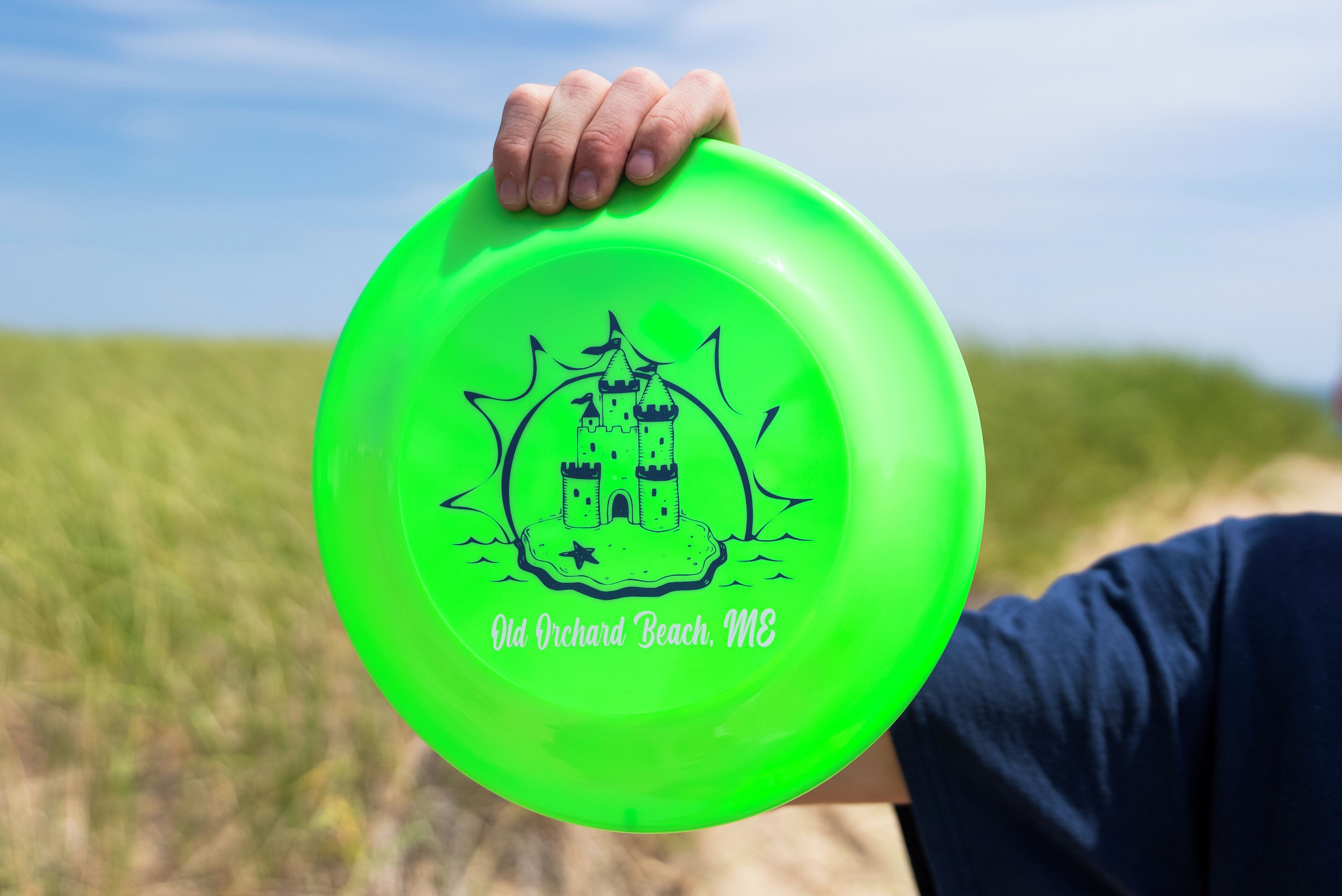 Een persoon houdt een felgroene frisbee vast, versierd met een kasteel ontwerp en de tekst 'Old Orchard Beach, ME'.