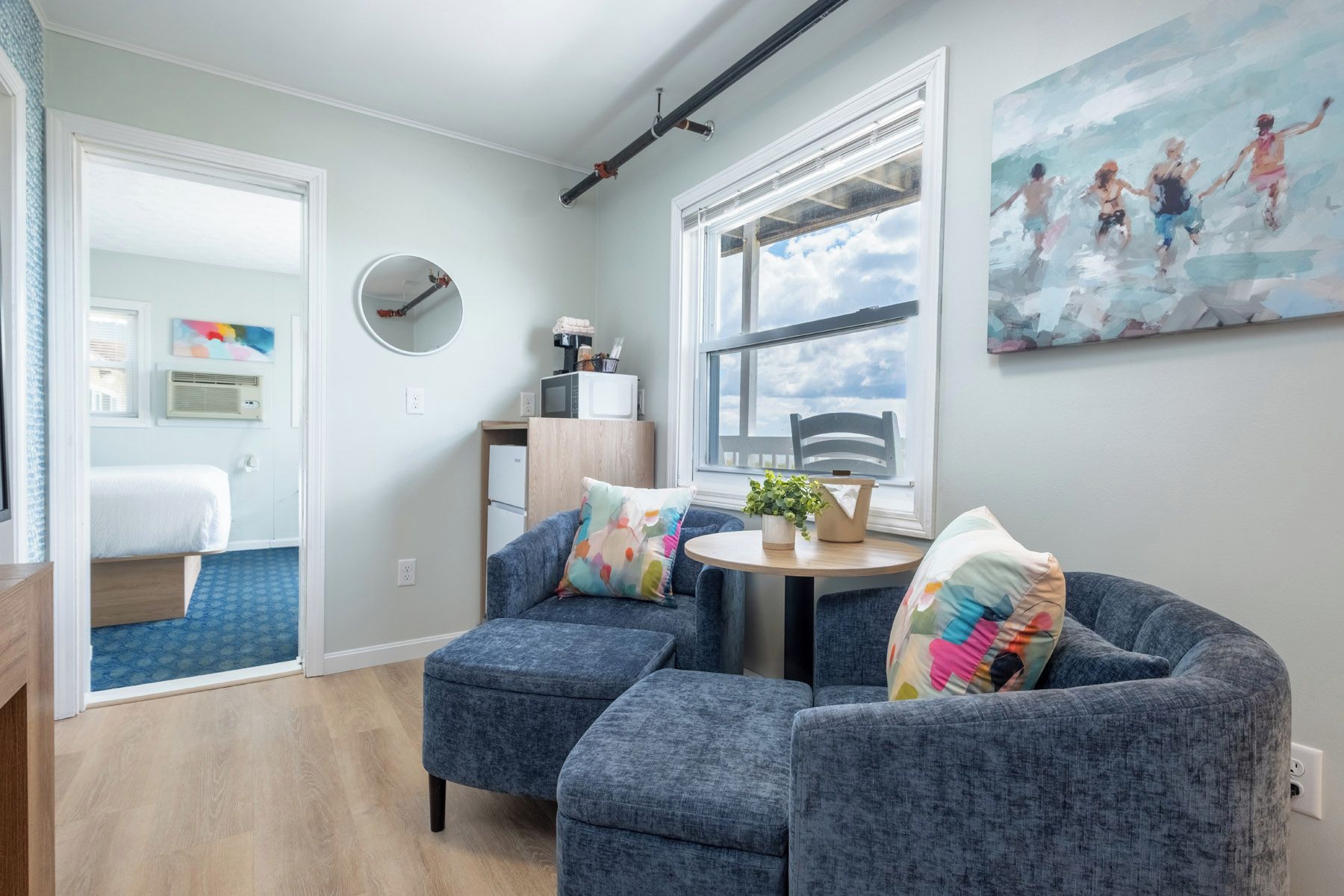 A modern living room with a round table, two blue armchairs, a large window with a view, and a painting on the wall.