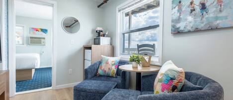A modern living room with a round table, two blue armchairs, a large window with a view, and a painting on the wall.