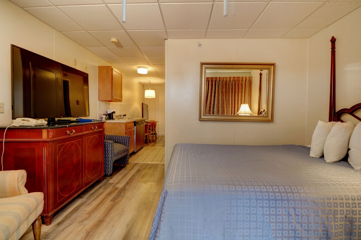 A hotel room with a large bed, a wooden dresser, a desk with a telephone, a chair, a small kitchenette, and a bathroom with a mirror and curtains.