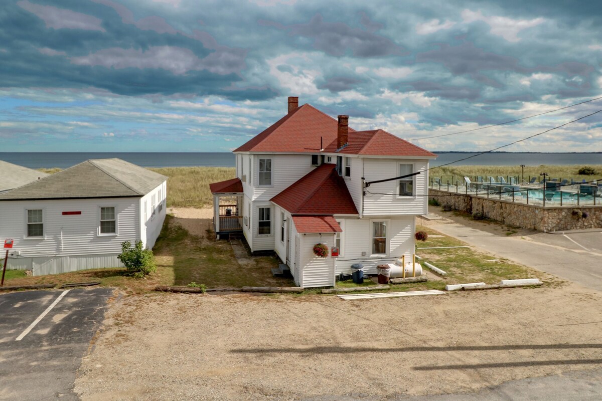 A two-story white house with a red roof, a small porch, and a view of the beach and ocean.