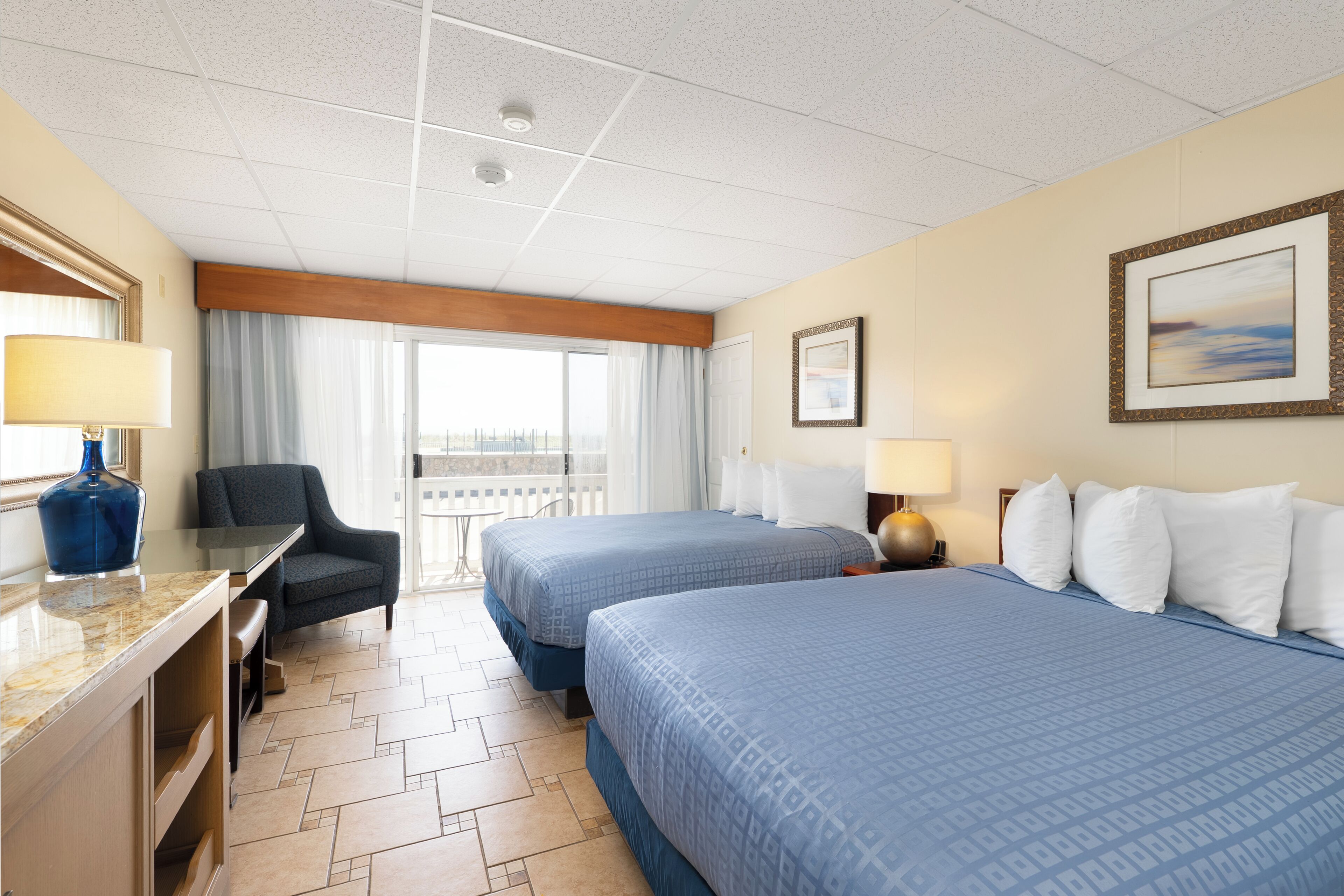 A hotel room with two beds, a desk, a chair, a window with curtains, and a framed picture on the wall.
