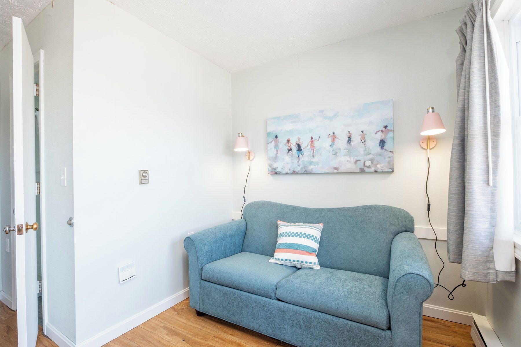 A room with a light blue sofa, a wall-mounted painting, two wall-mounted lamps, and a door.