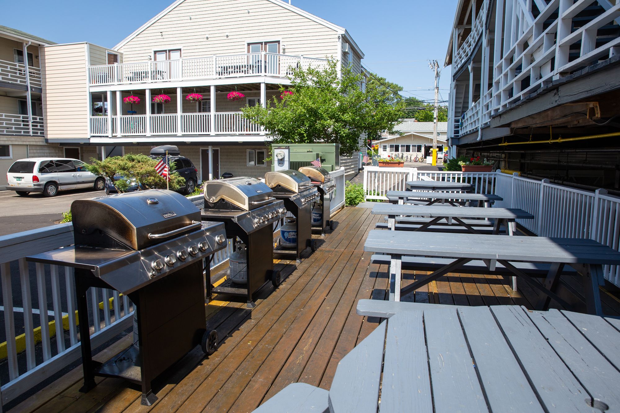 A deck with barbecue grills and picnic tables, overlooking a street with buildings and parked cars.
