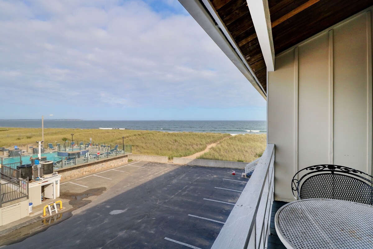 A balcony with a view of the beach and ocean, a parking area, and a table and chair.
