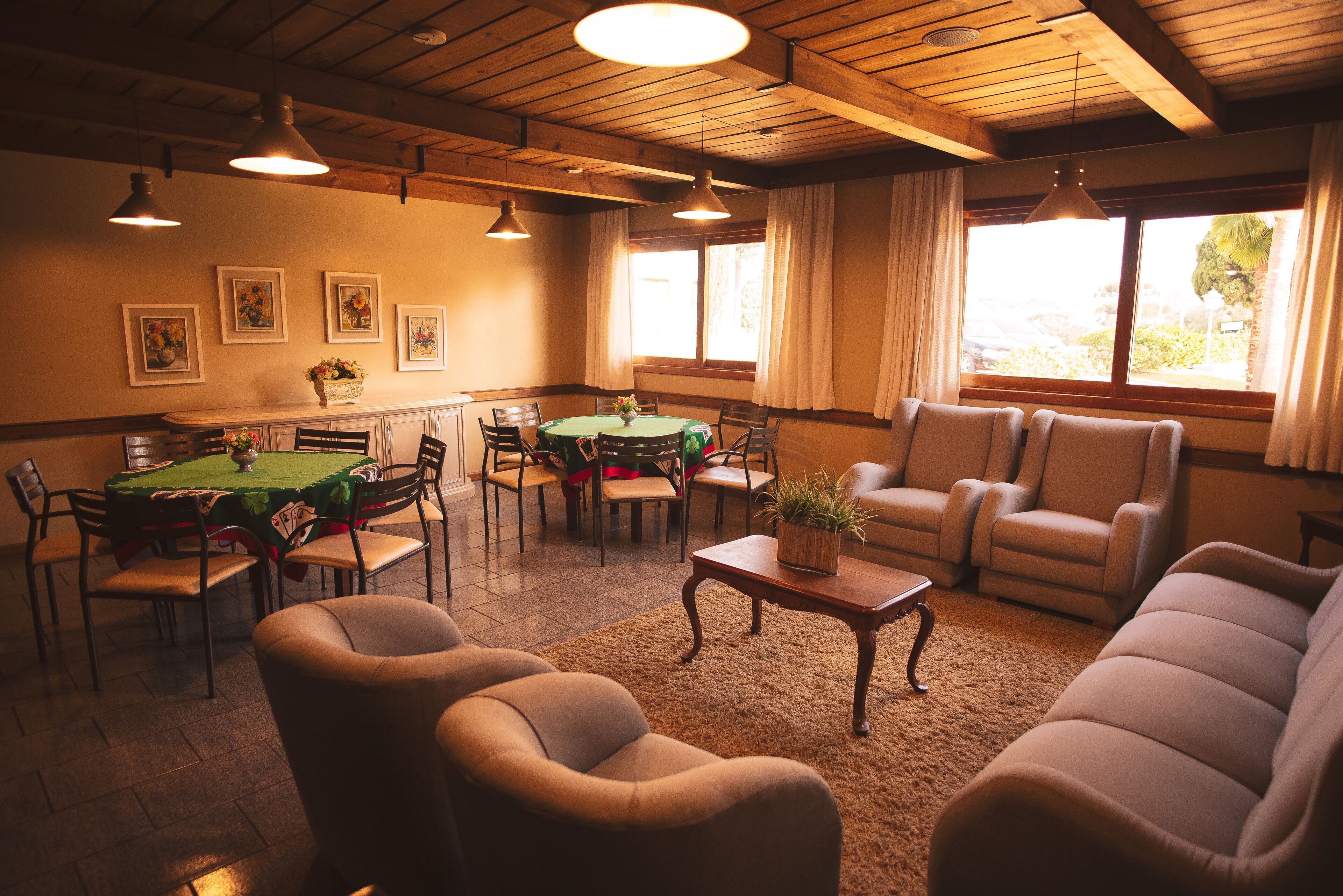 Uma sala de jantar com uma mesa redonda, cadeiras e toalha de mesa verde. Há quadros nas paredes, uma janela com cortinas e um teto com vigas de madeira.