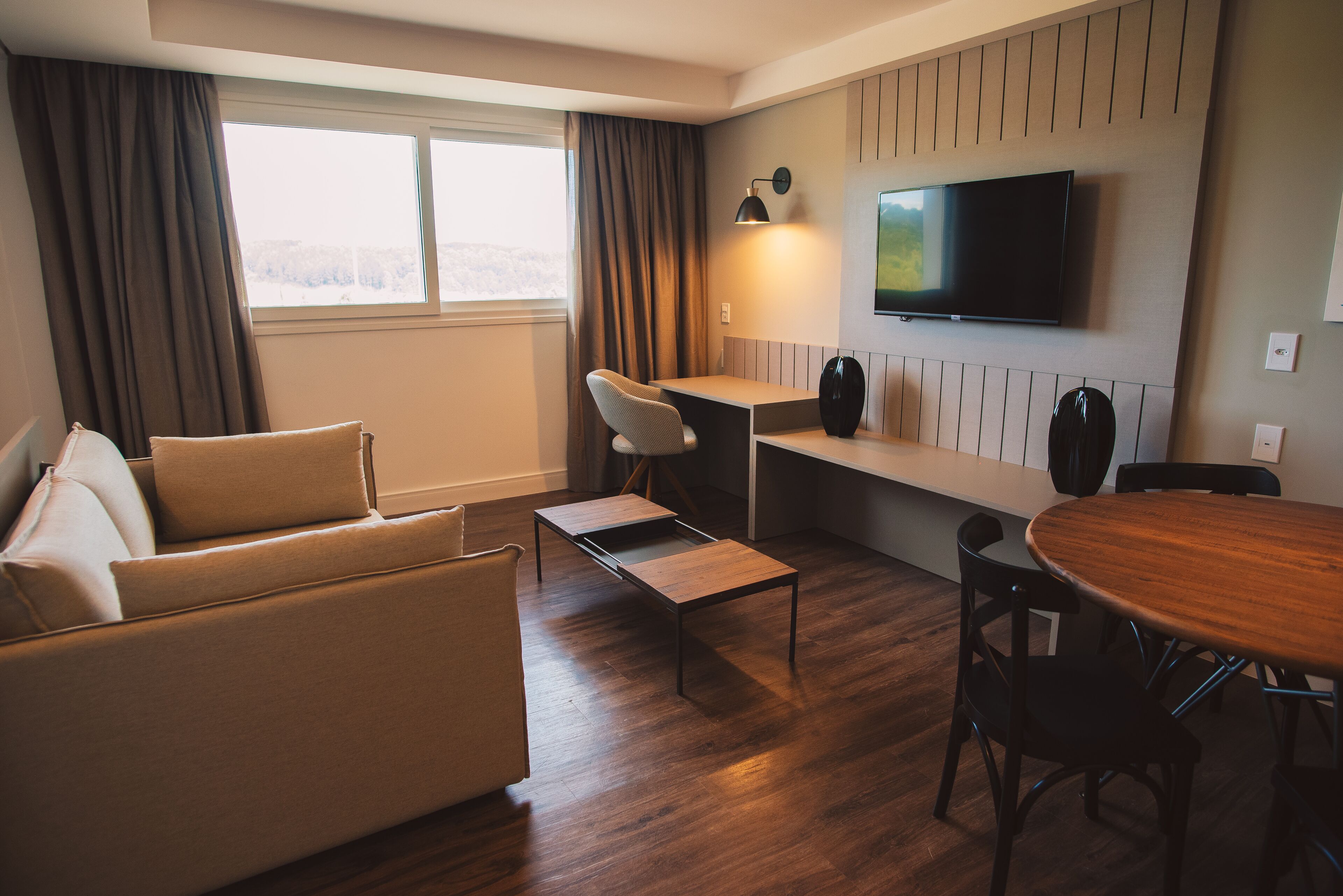 A modern hotel room with a sofa, a small desk, a dining table, and a TV mounted on the wall.