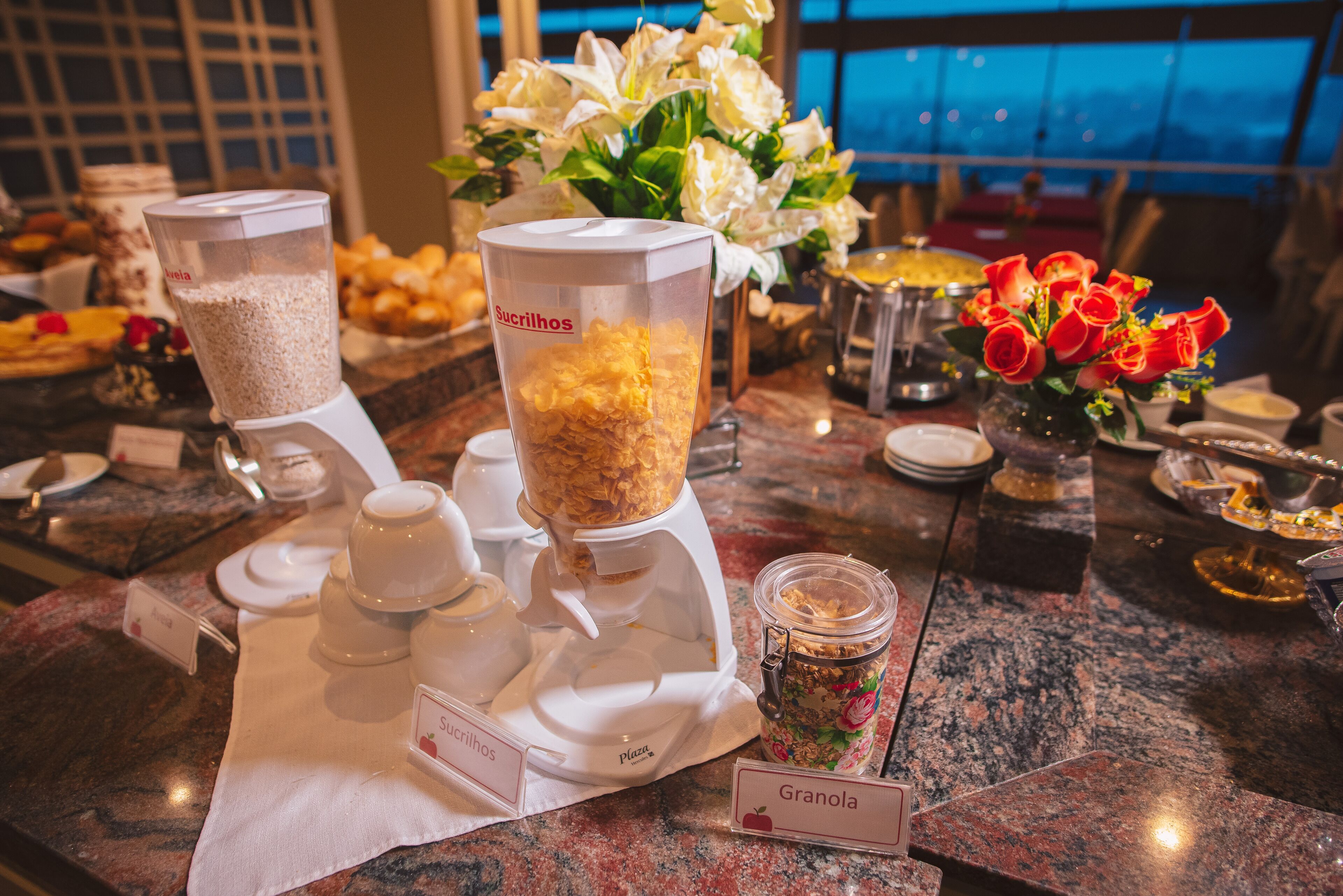 Montaje tipo bufé con estaciones de comida identificadas para sucralhos y granola, y un florero decorativo con rosas rojas.