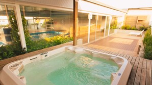 A hot tub on a wooden deck with a glass-enclosed patio area and a view of a swimming pool.