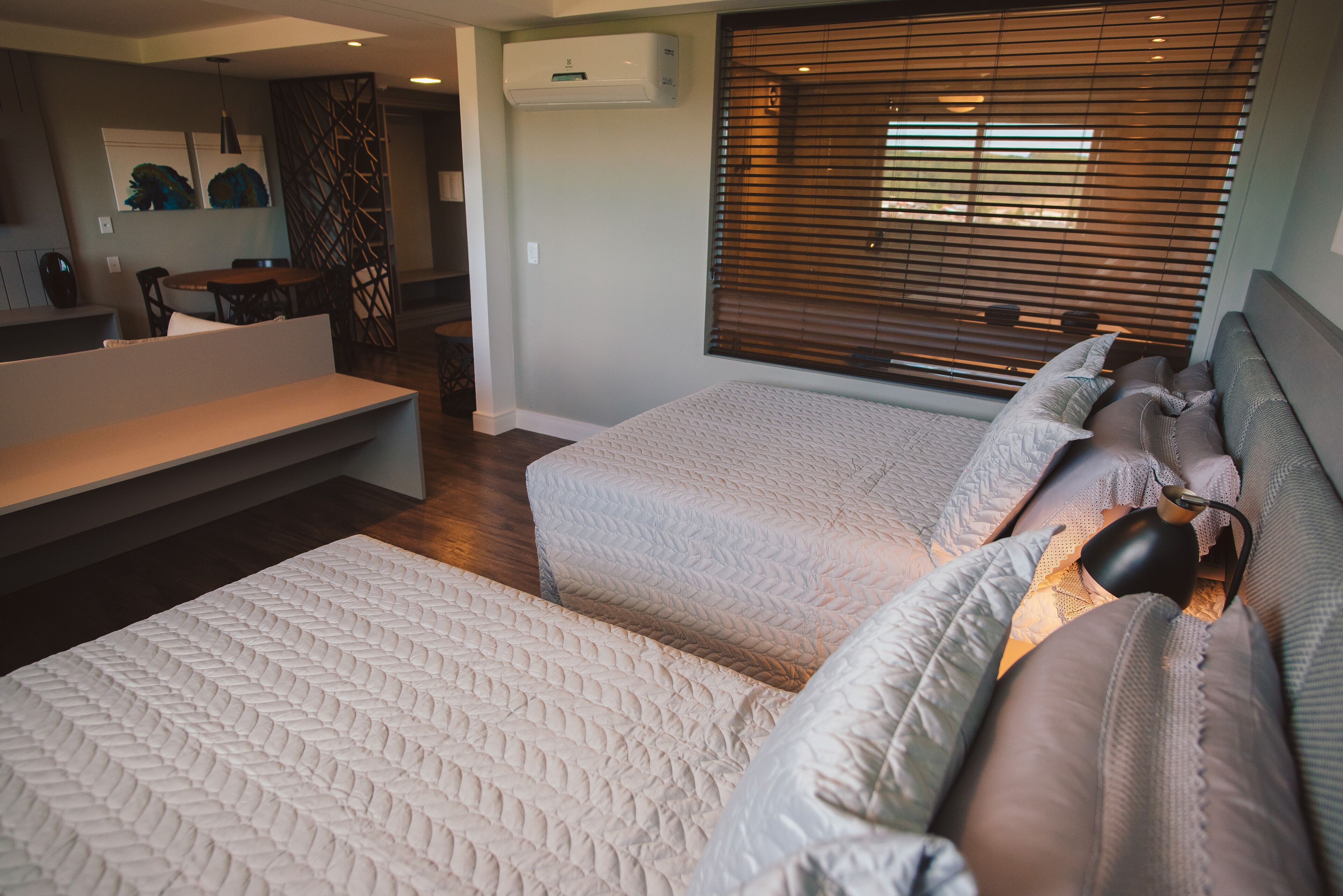 A modern bedroom with two beds, a sofa, a dining area, and a window with blinds.